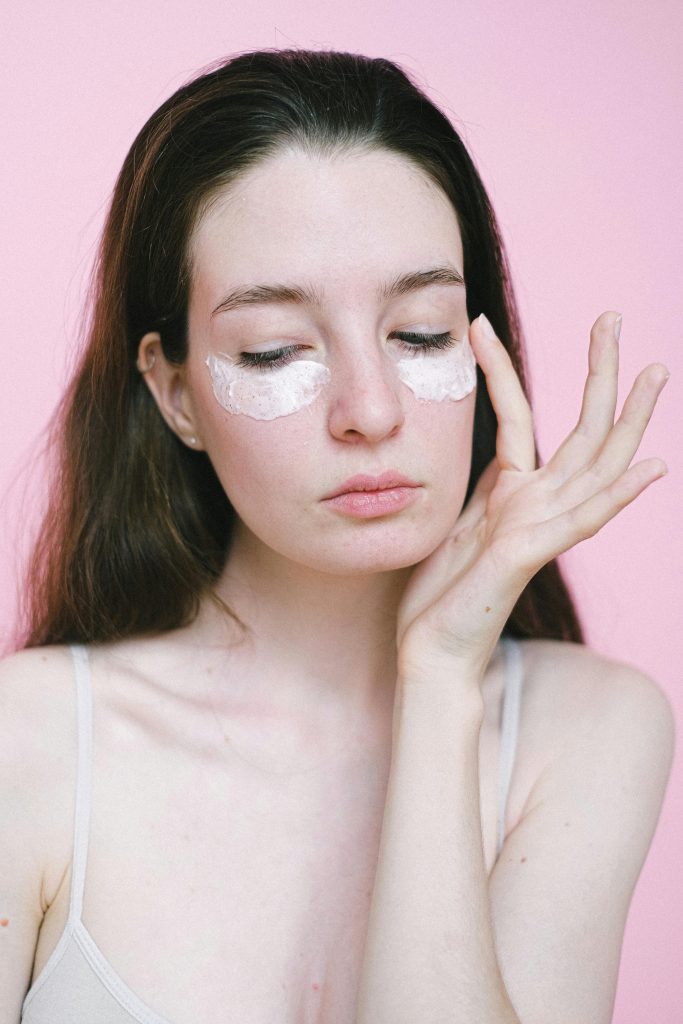 Jeune femme aux cheveux foncés appliquant une crème hydratante pour le visage sous les yeux sur un fond rose en studio